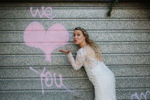 Bride in lace dress poses playfully with pink heart graffiti on wall.
