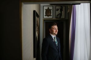 Groom in dark suit stands by window with dramatic lighting and wall art.