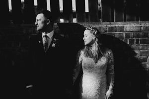 Black and white wedding photo of a couple by a brick wall in an artistic composition.