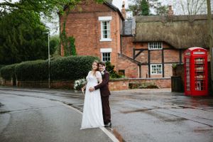 Wedding couple poses in front of brick building and classic red British phone booth on rainy day.