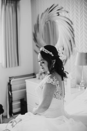 Bride in lace dress sits contemplatively beside ornate mirror in bridal suite.