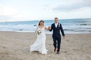 Couple walking hand in hand on a beach in wedding attire with ocean waves in the background.