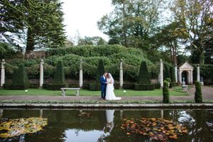 Wedding couple stands by a reflecting pool in a formal garden with classical architecture and fall foliage.
