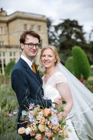 Newlyweds pose in garden with colorful bouquet against historic manor house.