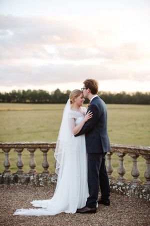 Romantic wedding couple embracing at sunset on a balcony overlooking countryside grounds.