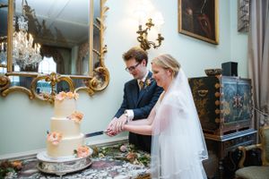 Wedding couple cutting their elegant peach-colored cake in ornate room with gilded mirrors.