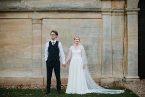 A wedding couple poses against a stone wall, the bride in a flowing white dress and the groom in formal attire.