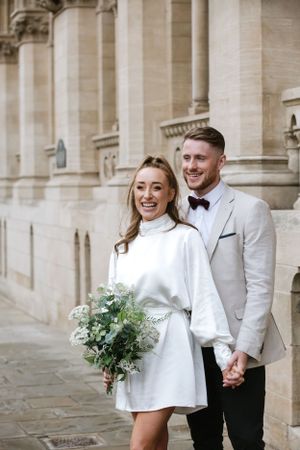A stylish couple in white formal wear poses against classic stone architecture.