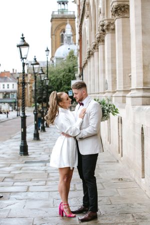 Elegant couple in wedding attire embracing on historic street with classical architecture and lampposts.