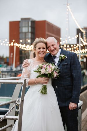 Wedding couple posing on boat dock with string lights and urban backdrop at dusk.