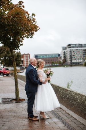 Wedding couple standing by waterfront with autumn tree and cityscape in background.