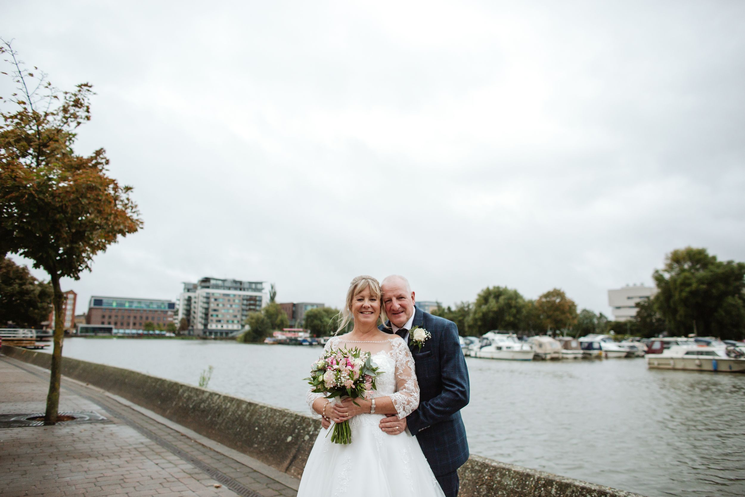 Newlyweds share a joyful moment by the waterfront with boats and autumn trees in the background.