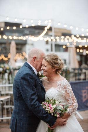 Romantic evening wedding portrait under twinkling string lights with bride holding a pink and green bouquet.