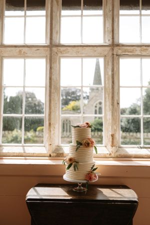 Three-tiered white wedding cake with floral decorations displayed against a bright window with garden views.