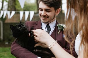 Wedding guest in burgundy suit holding black cat during outdoor reception.
