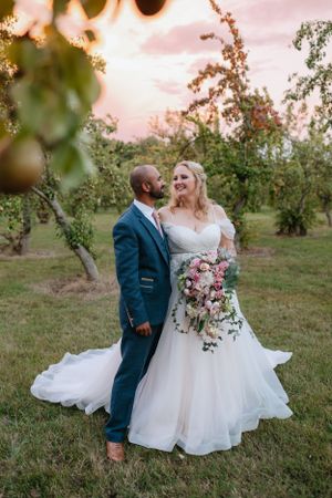 Newlyweds share a joyful moment together in an orchard at sunset with a flowing white dress and navy suit.