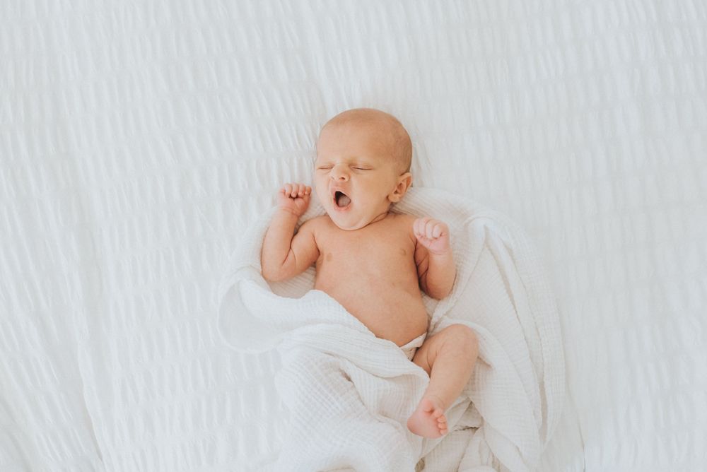 Sleeping newborn baby in white lying on soft white bedding with outstretched arms.