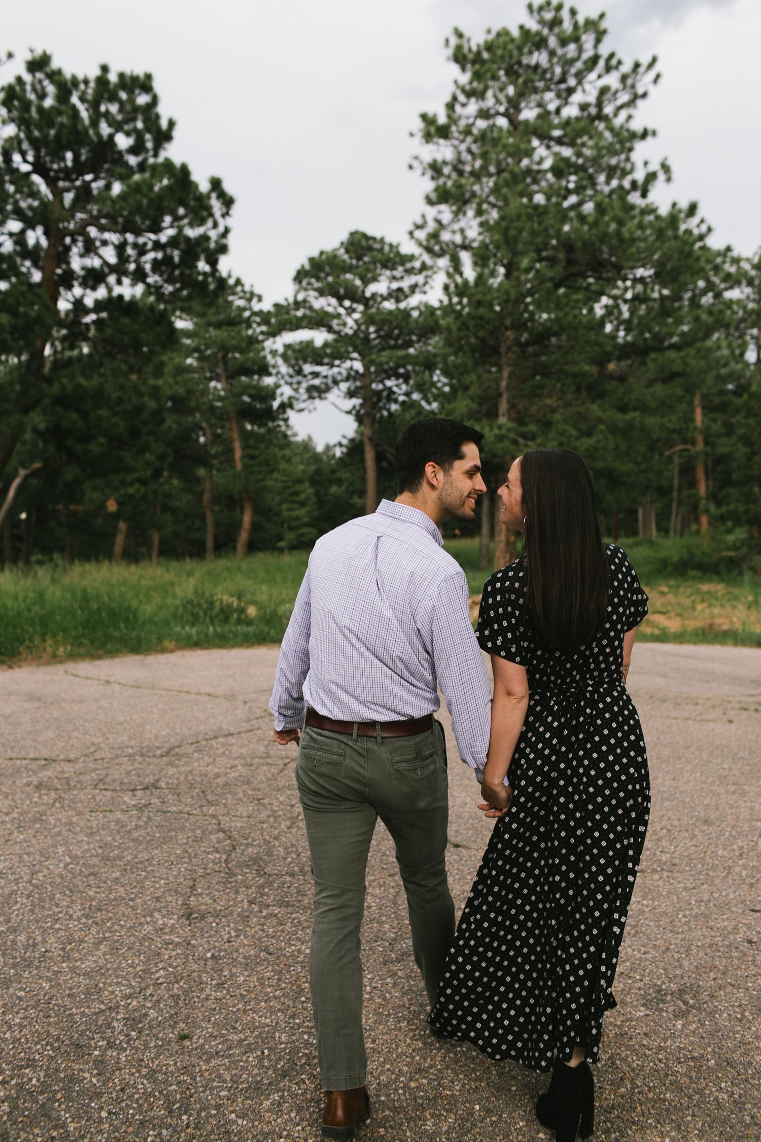 Couple in polka dot dress and striped shirt walking together on a path near pine trees.