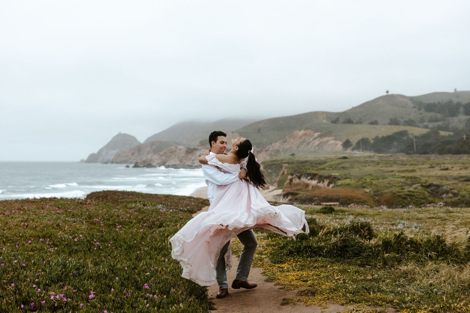 man twirling his girlfriend around with the mountains and ocean and beach in the background in Pacifica near SF Bay Area, California
