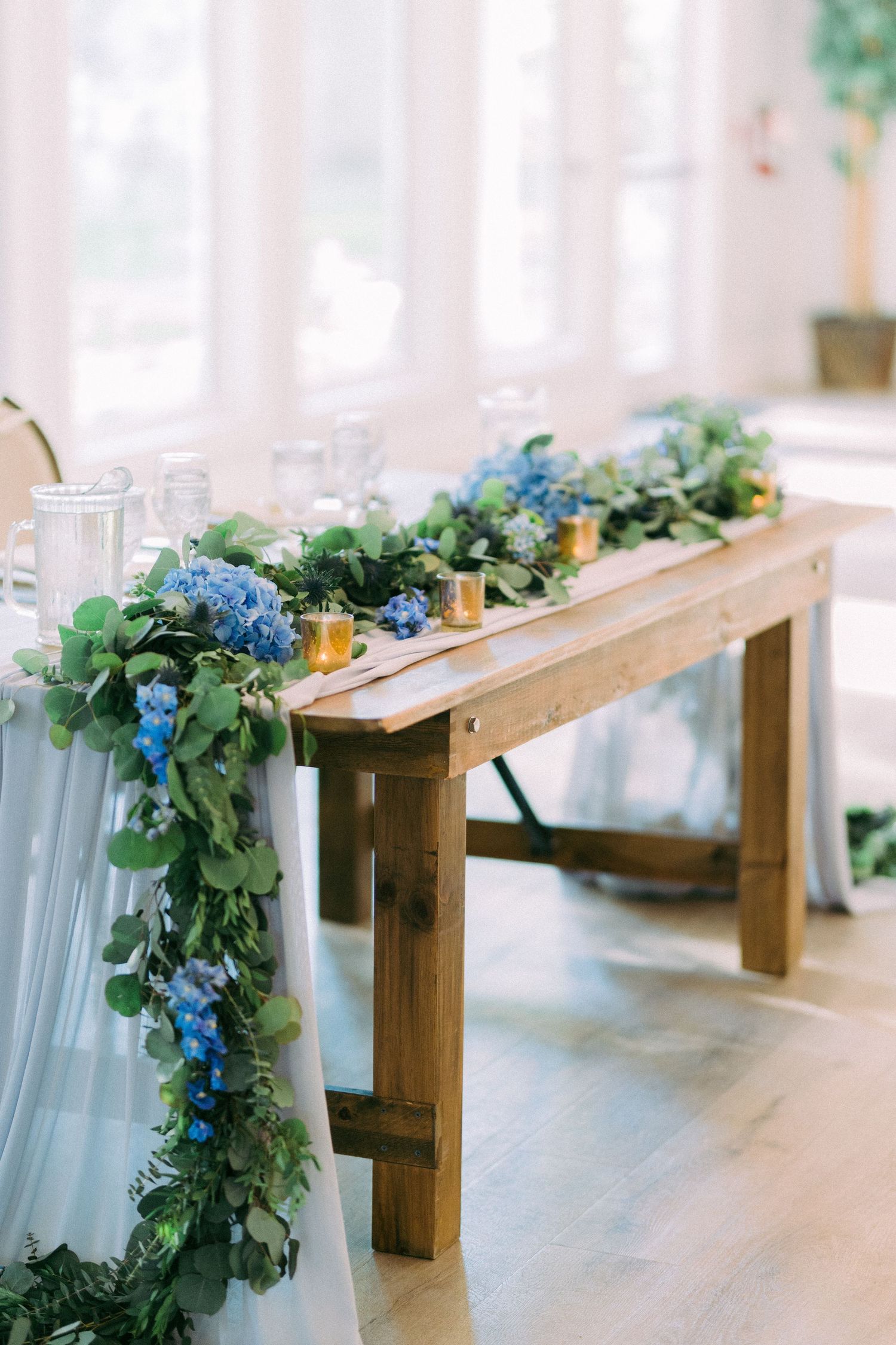 Rustic wooden bench decorated with blue hydrangea garland and flowing fabric for wedding decor.