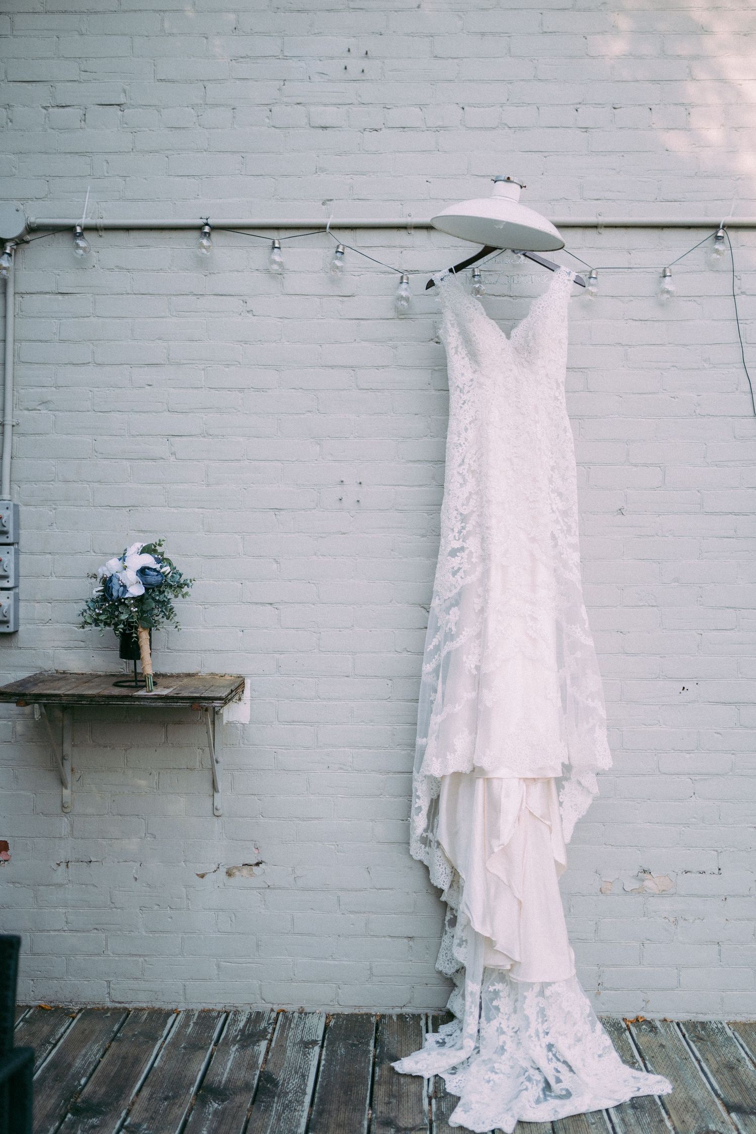 Elegant white lace wedding dress hanging on brick wall with blue floral bouquet on rustic wooden shelf.
