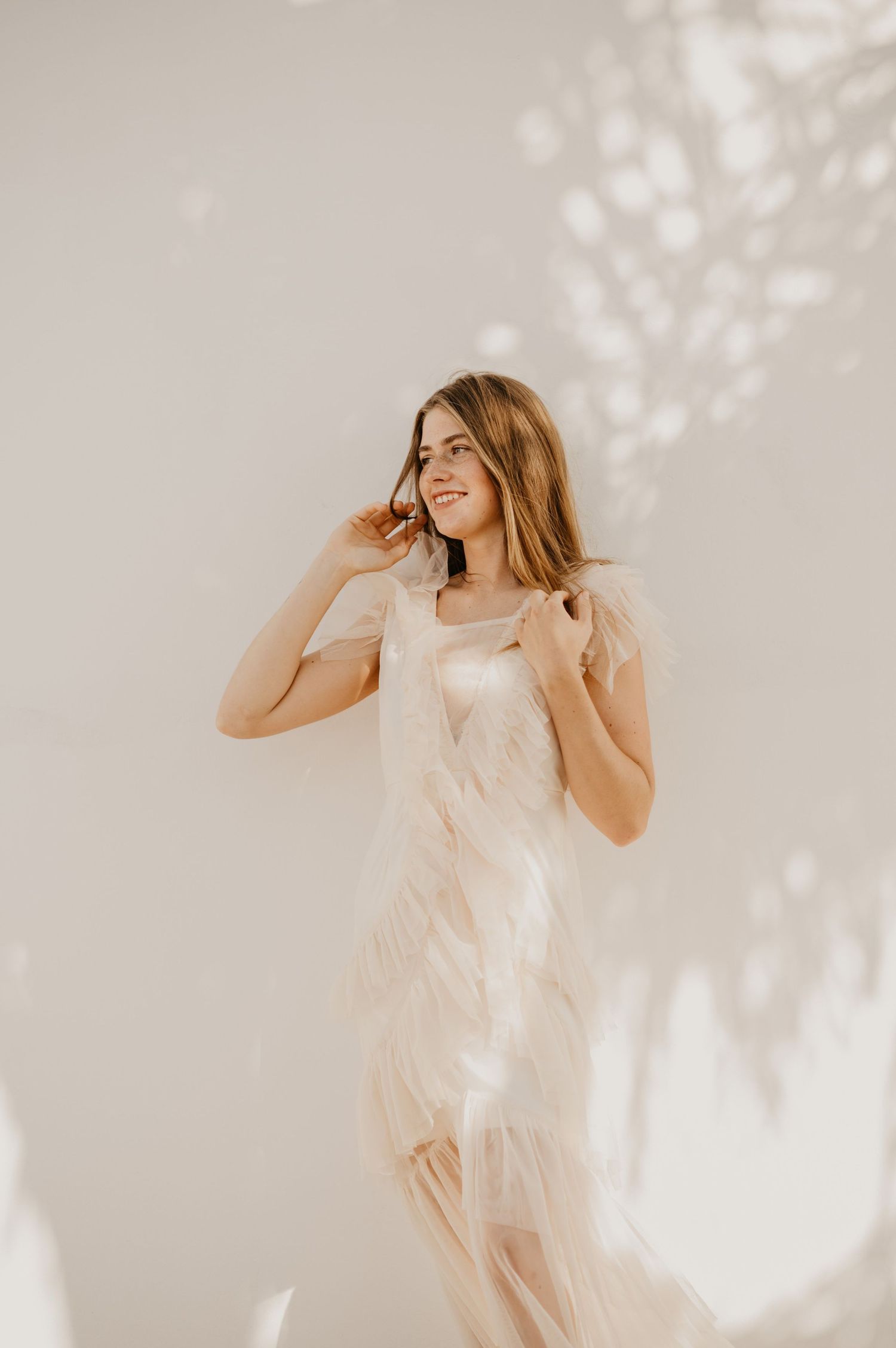 A smiling woman in a flowing blush feathered gown poses gracefully against a soft white wall with dappled light.