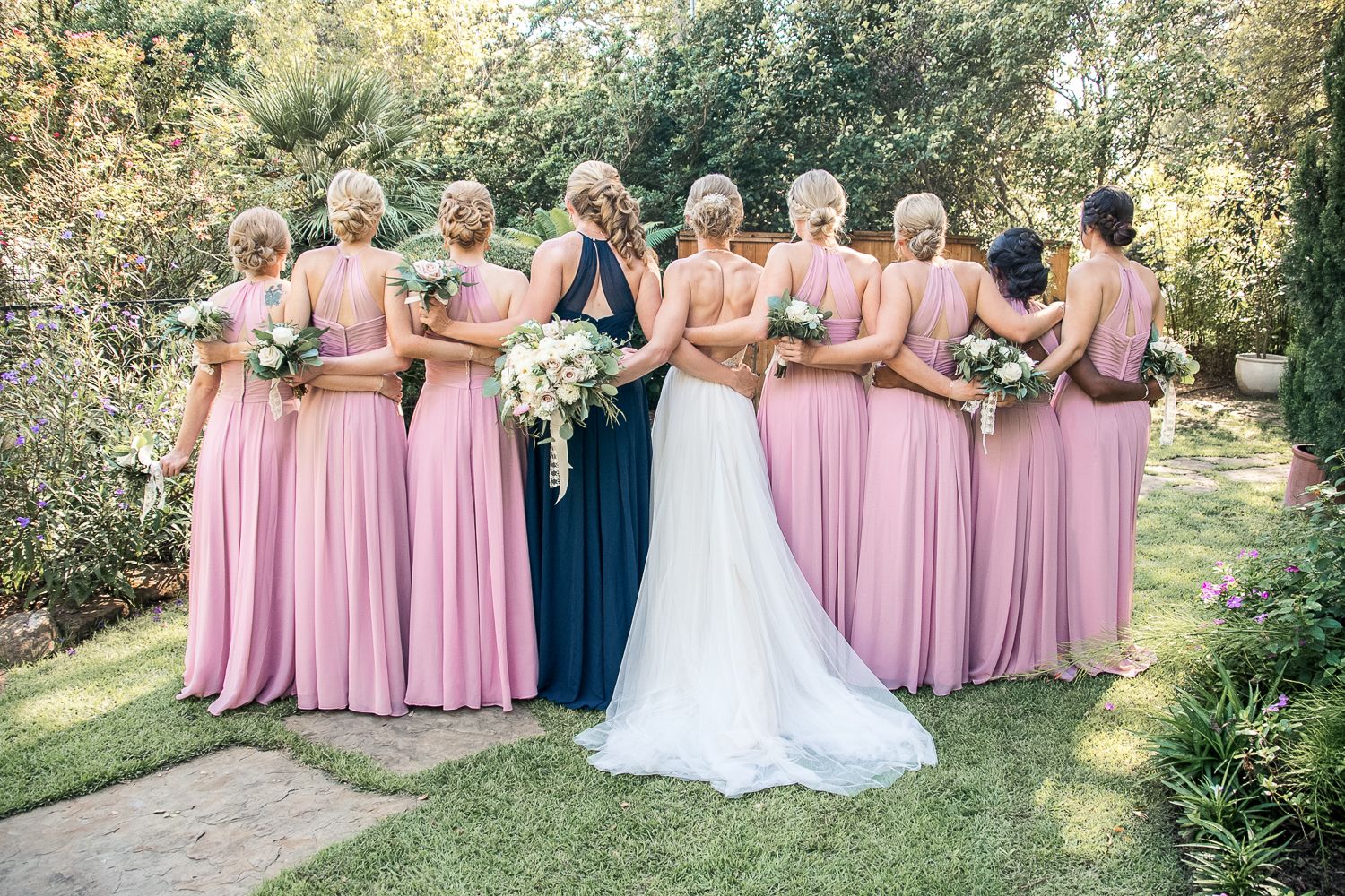 Wedding party in pink dresses poses together outdoors on garden pathway.