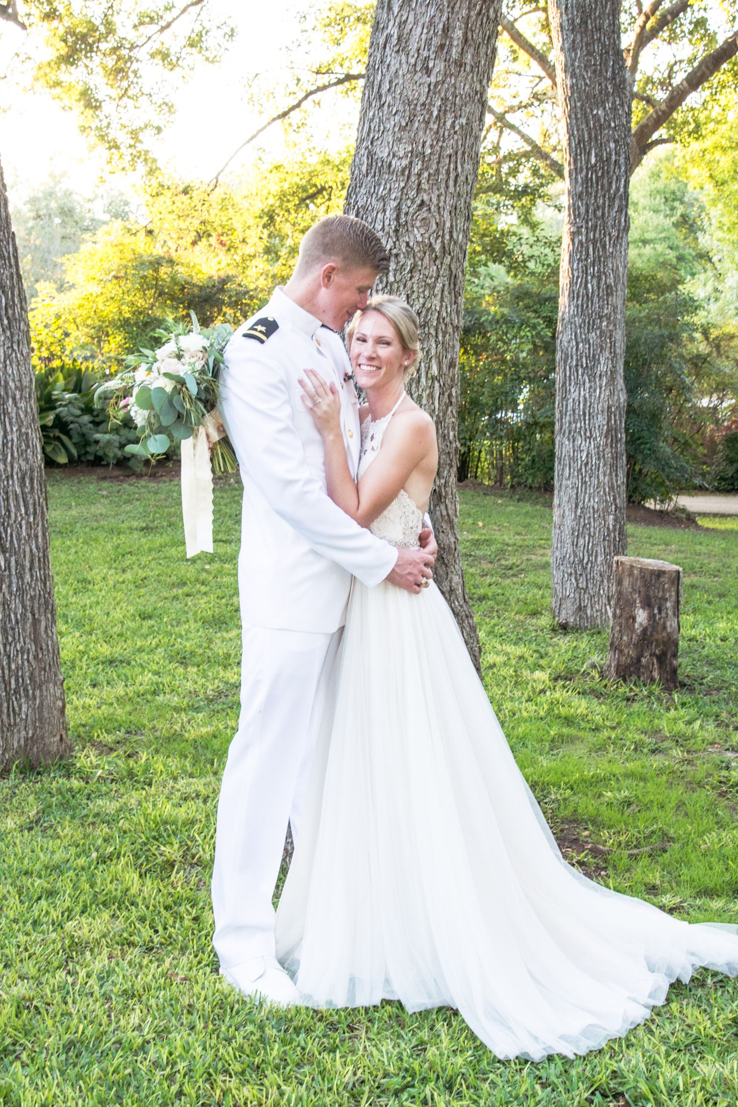 Elegant couple in white attire embraces on green lawn with tall trees in background.