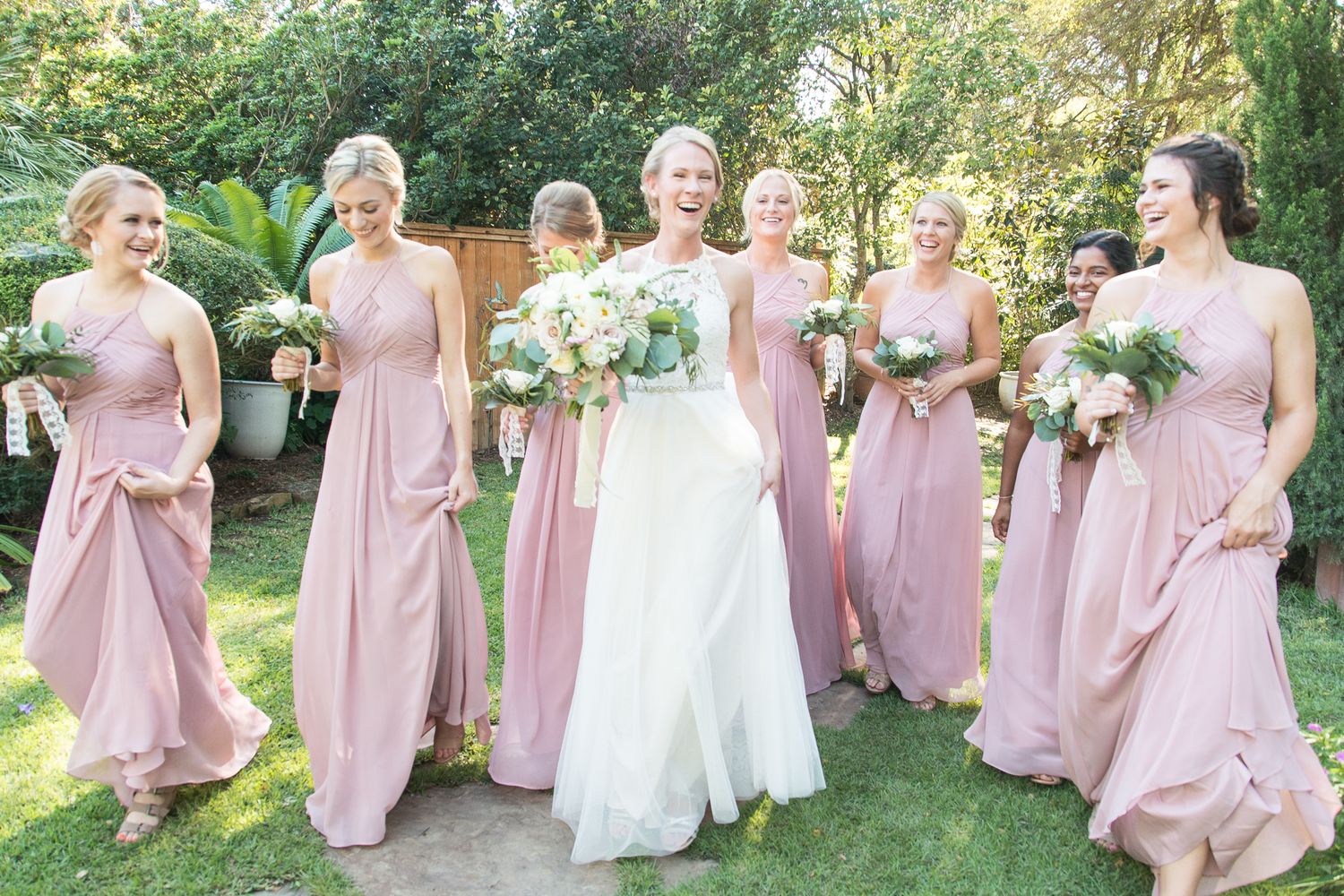Bridal party in dusty rose gowns poses on garden path with bouquets.