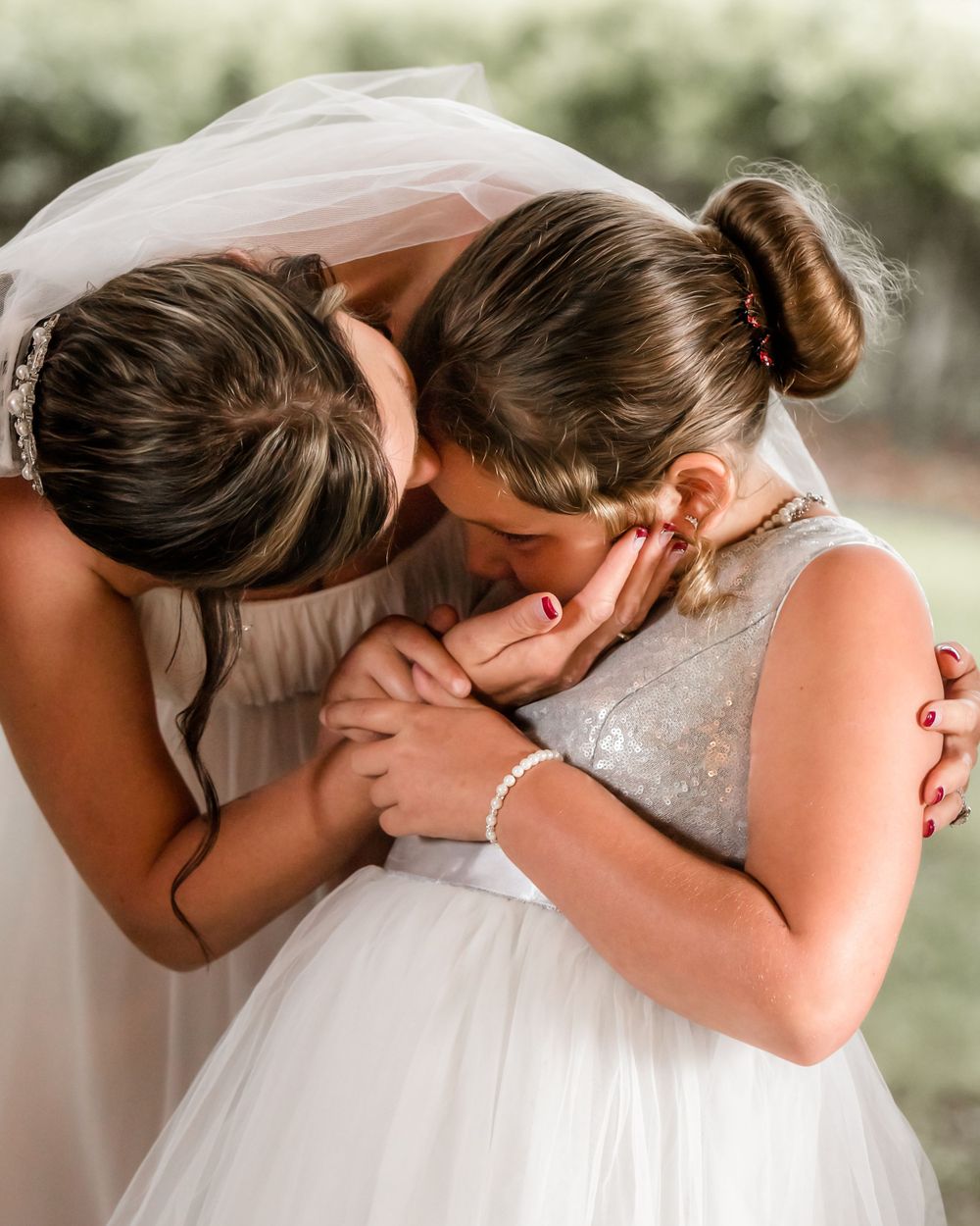 Two people in wedding attire share a tender embrace while one kisses the other's hands in an emotional outdoor moment.