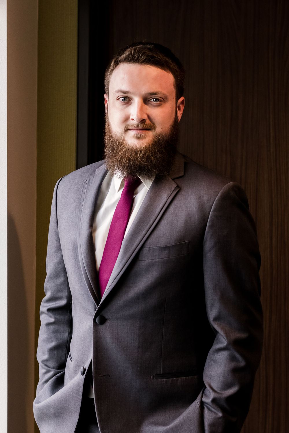 Professional portrait of a person in a gray suit and burgundy tie standing against a dark background.