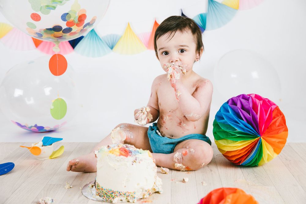 Baby wearing blue diaper smashing into birthday cake next to colorful decorations.