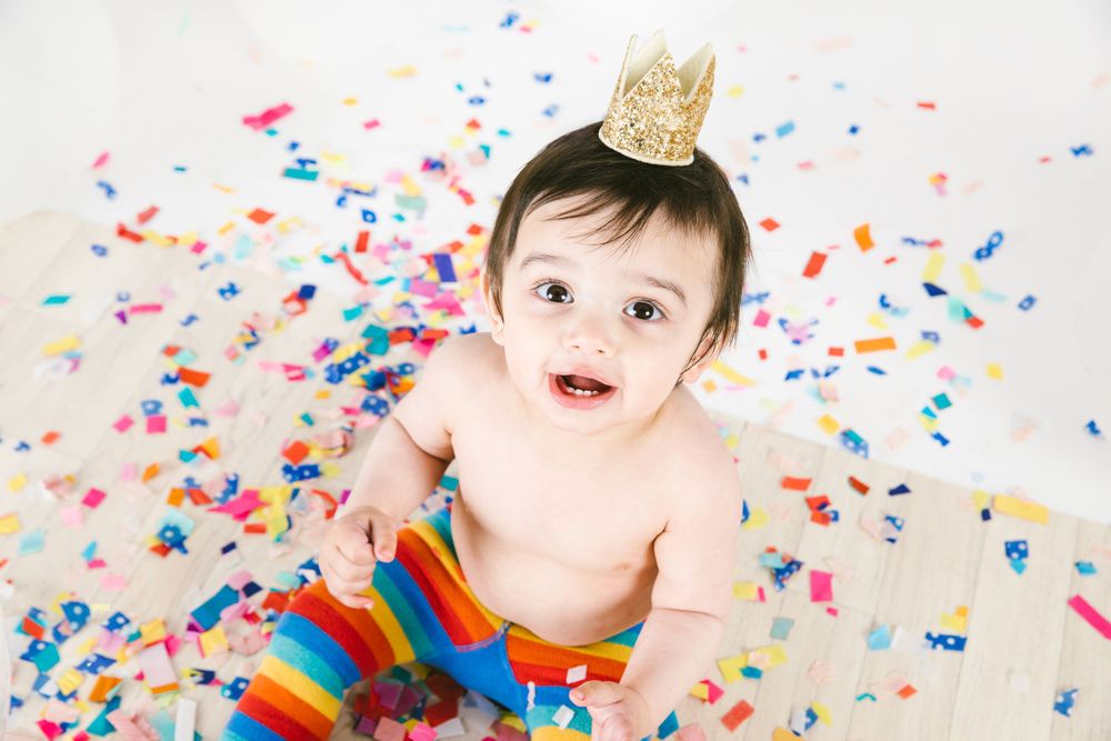 Baby wearing crown sits among colorful confetti with rainbow leg warmers.