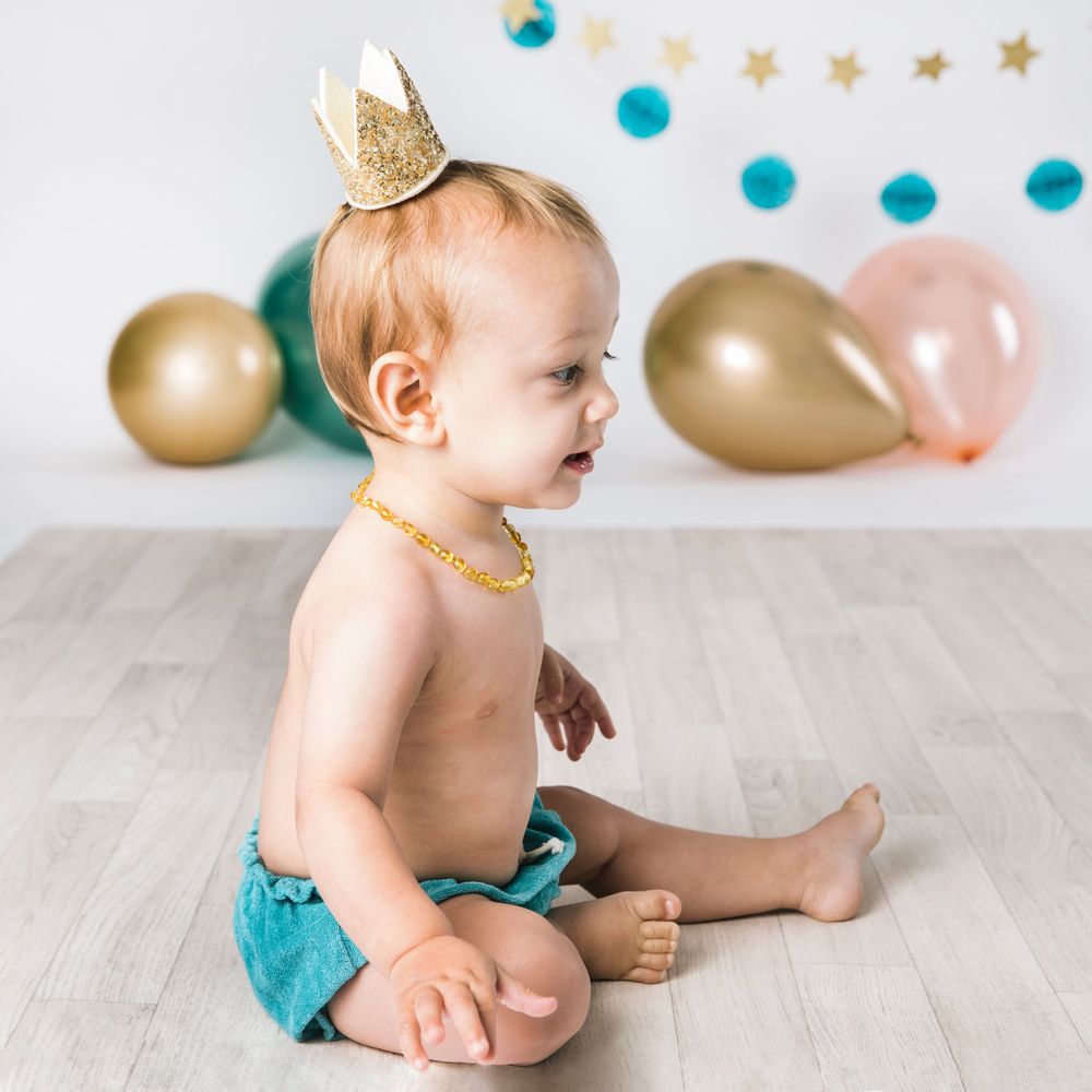 A baby wearing a tiny gold crown sits on a wooden floor with party decorations in the background.