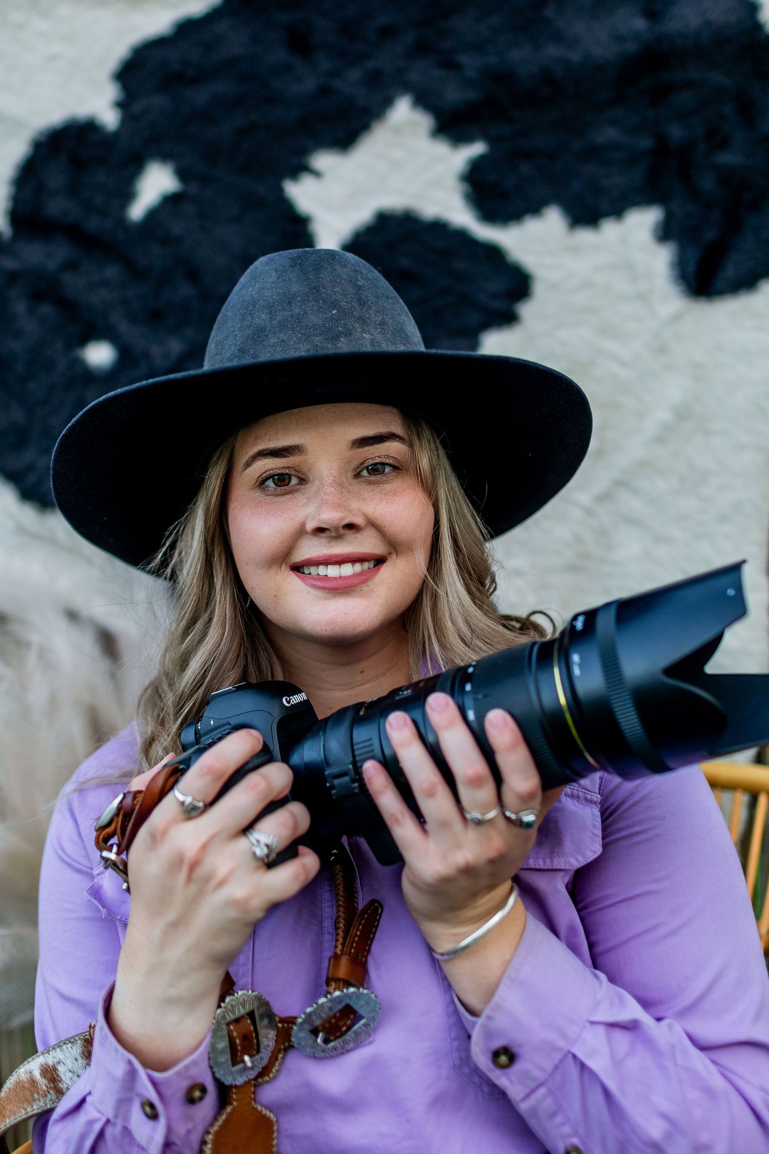Western style photographer in purple shirt and wide brim hat holds professional camera equipment.