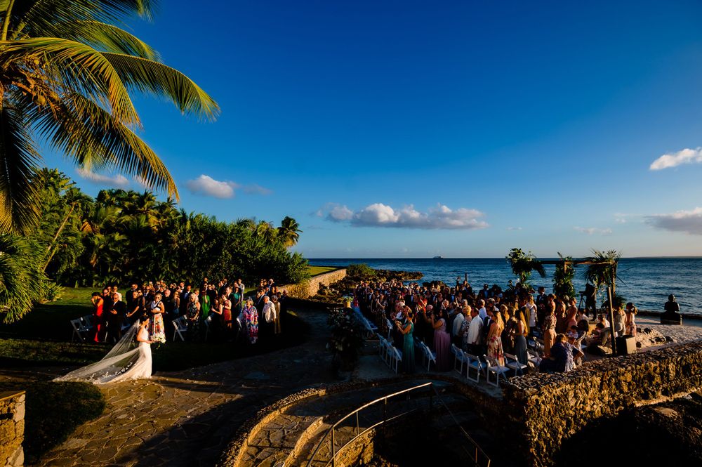 Bride walking down the aisle at a sunny beachside destination wedding ceremony.