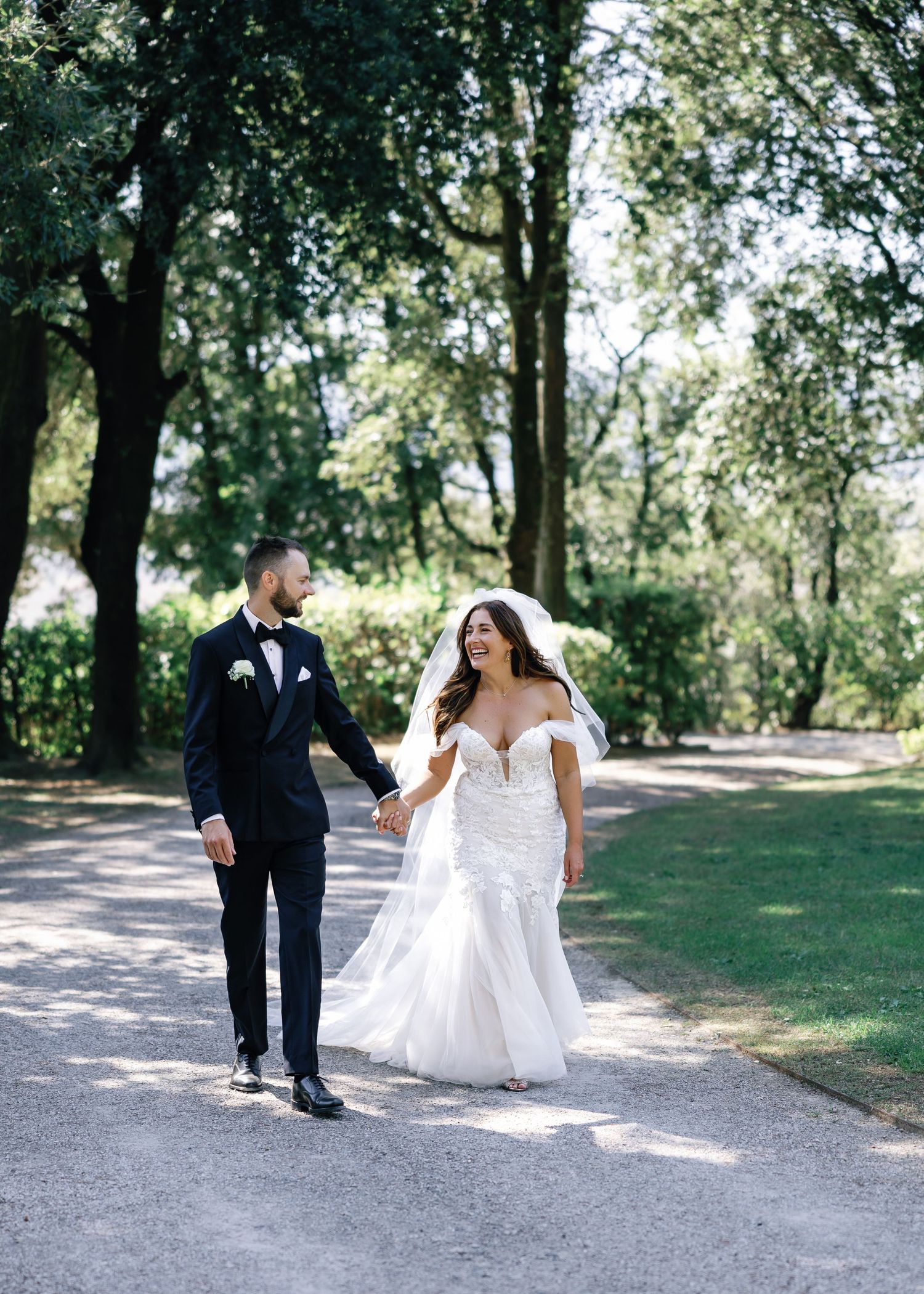 Newlywed couple walking along a tree-lined path on their wedding day in Italy with bride in flowing white gown.