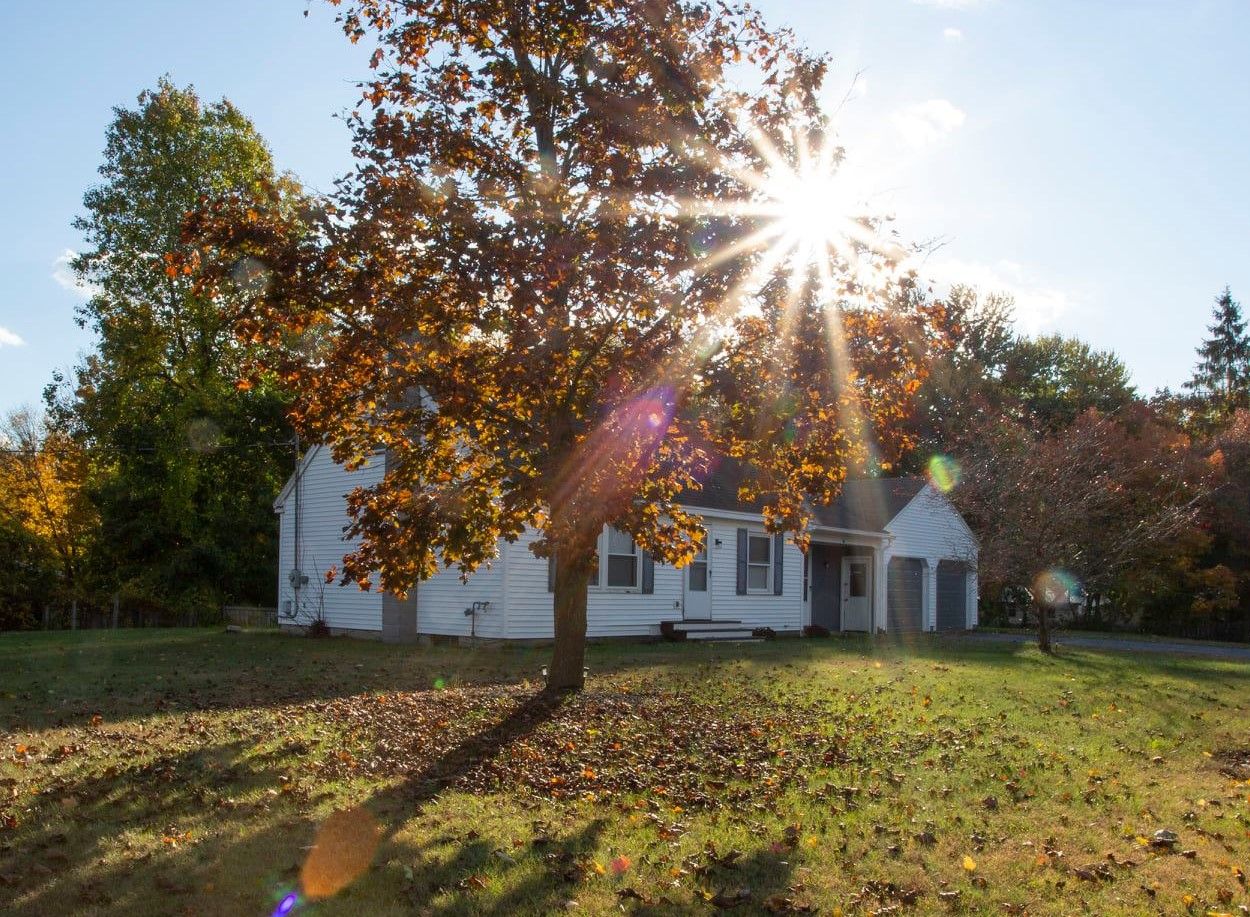 Sunburst through autumn trees illuminates white house and fallen leaves on grass.