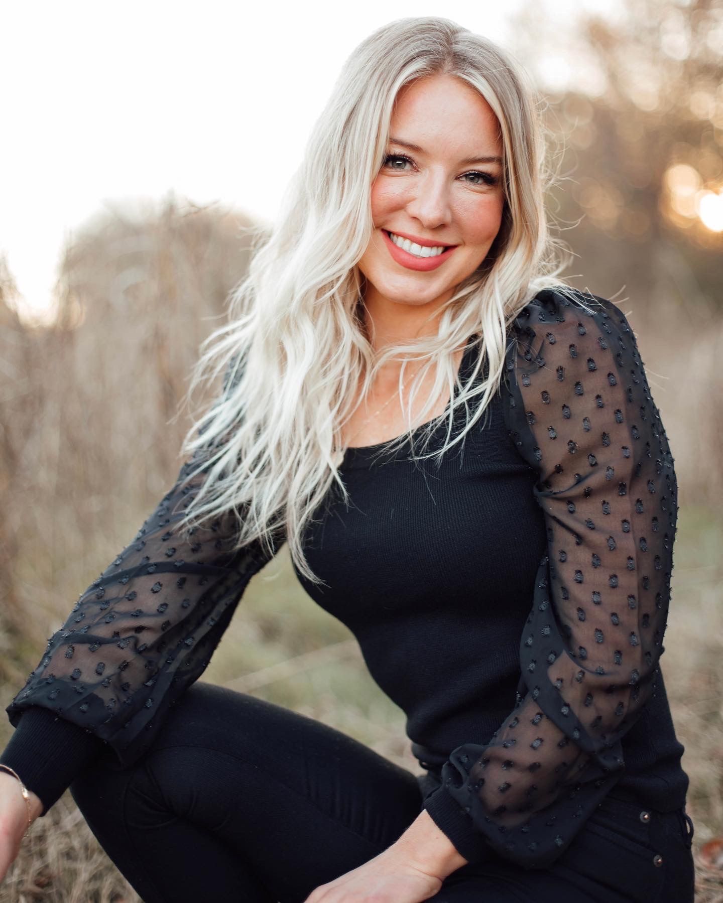 Woman in black top with sheer puff sleeves smiling outdoors at sunset.