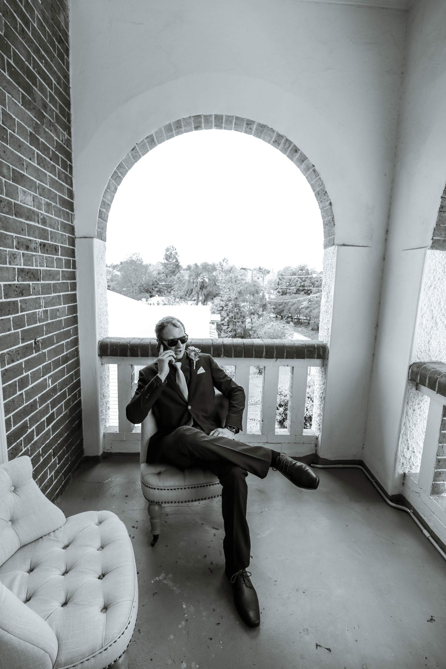 A stylish figure lounging in a black suit on a porch chair under an arched brick doorway.