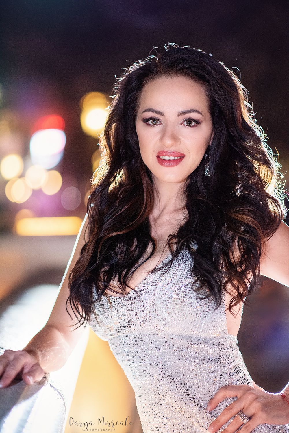 Woman in sparkly silver dress poses against nighttime city lights.