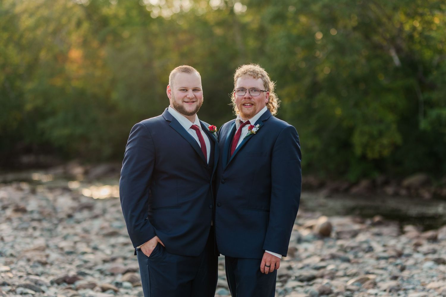 Two people in navy suits stand together outdoors on rocky terrain at sunset during a wedding celebration.