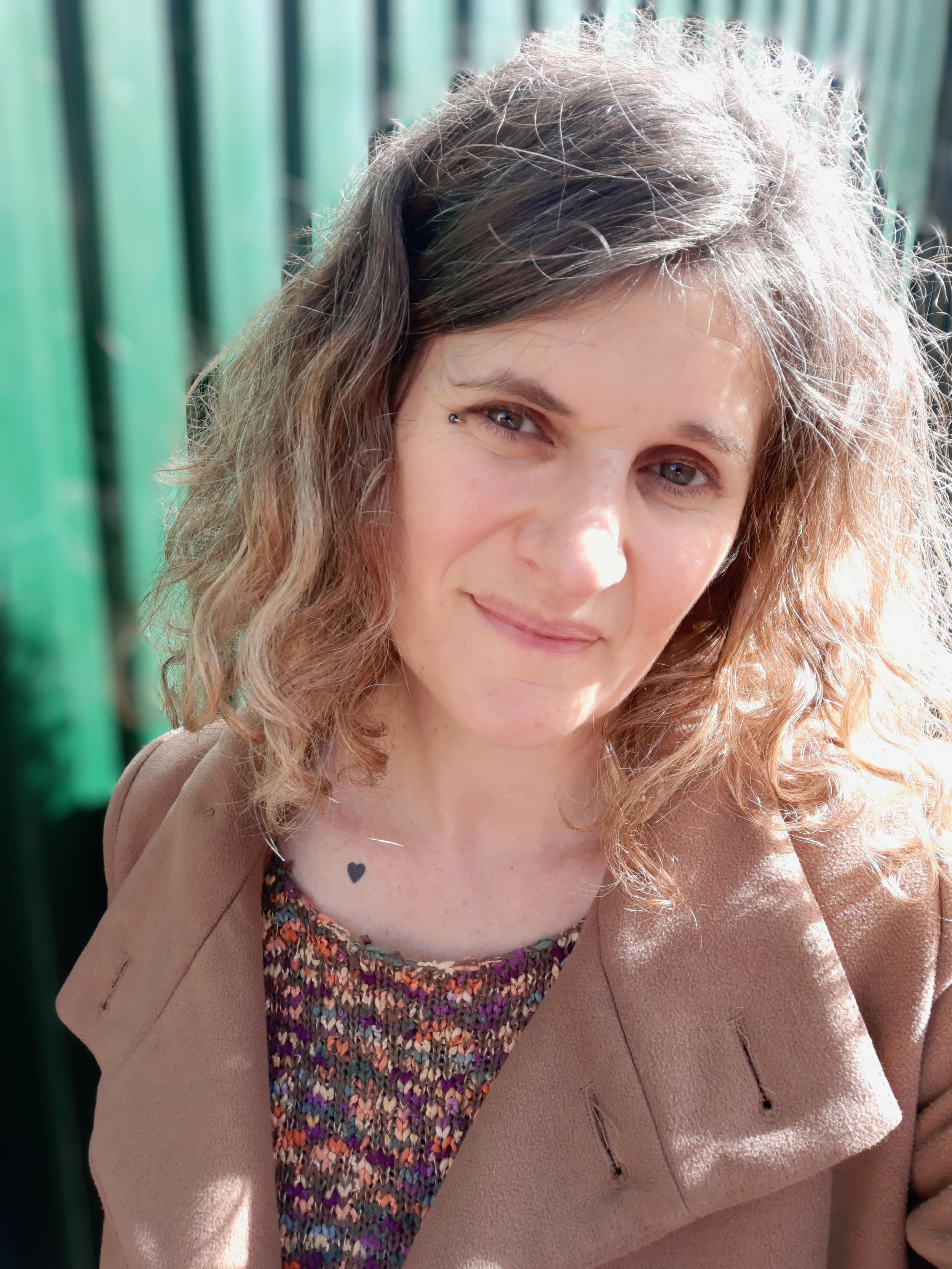 Close-up portrait with curly hair wearing floral top and tan blazer against green wall.