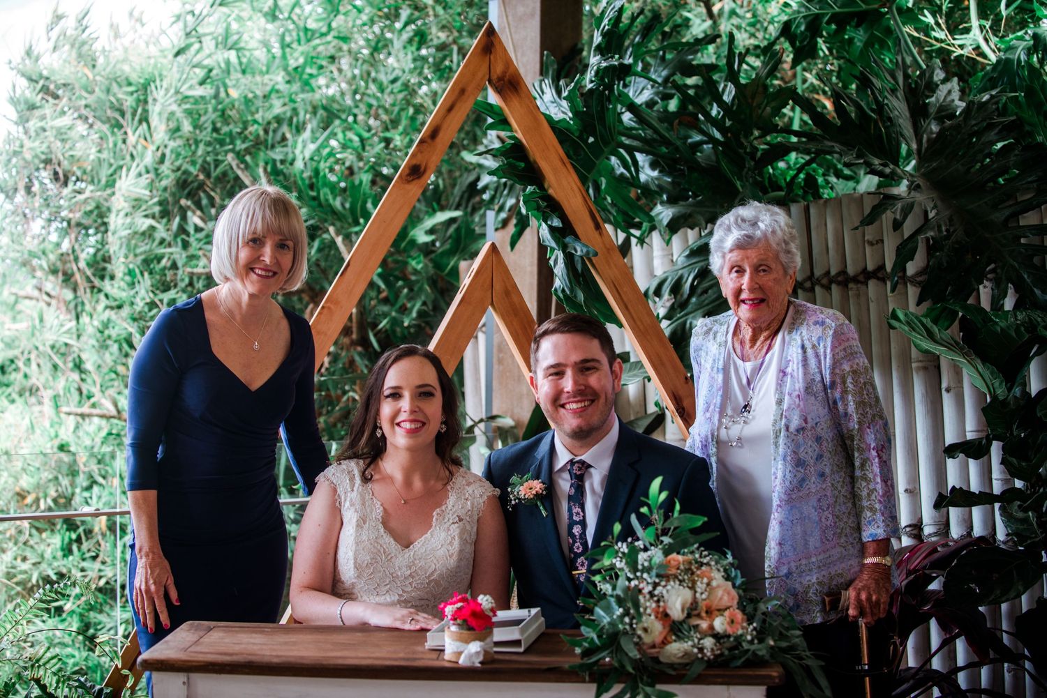 Group photo at wedding ceremony under wooden triangular arch with tropical decor.