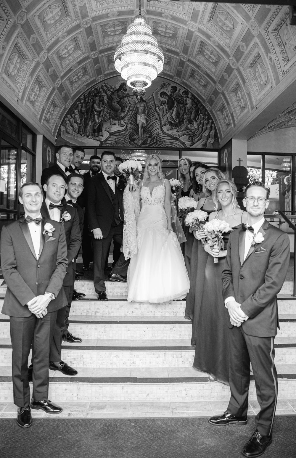 Wedding party stands elegantly on grand staircase with ornate chandelier and architectural details.