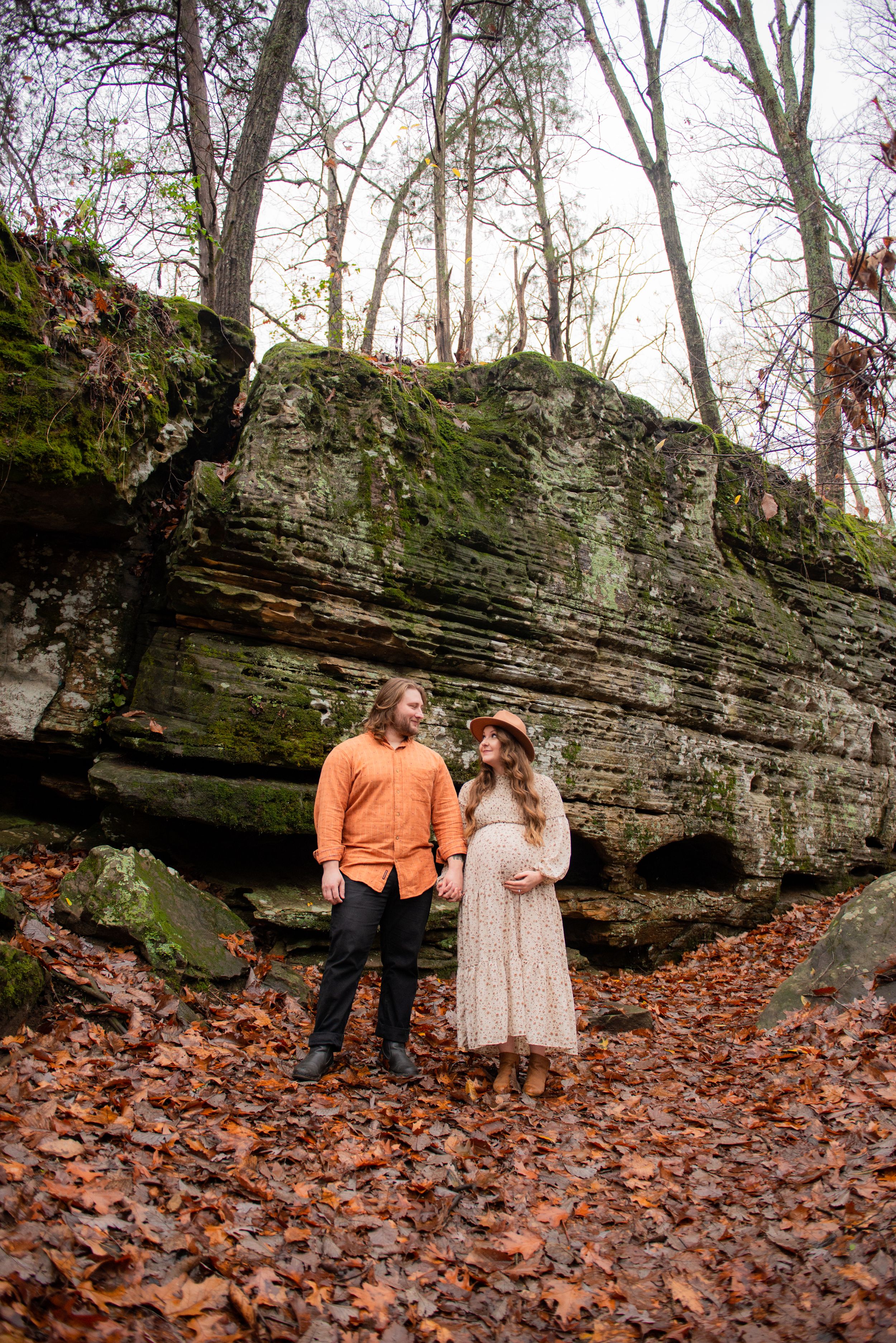 A couple poses near a rocky cliff surrounded by fallen autumn leaves.