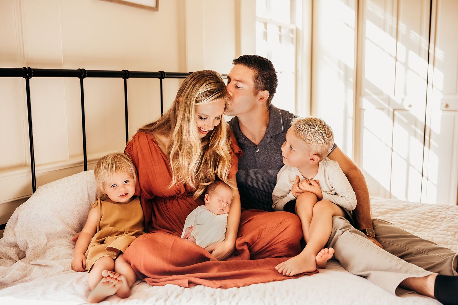 A family of 5 sits on their bed snuggling their newborn baby girl.