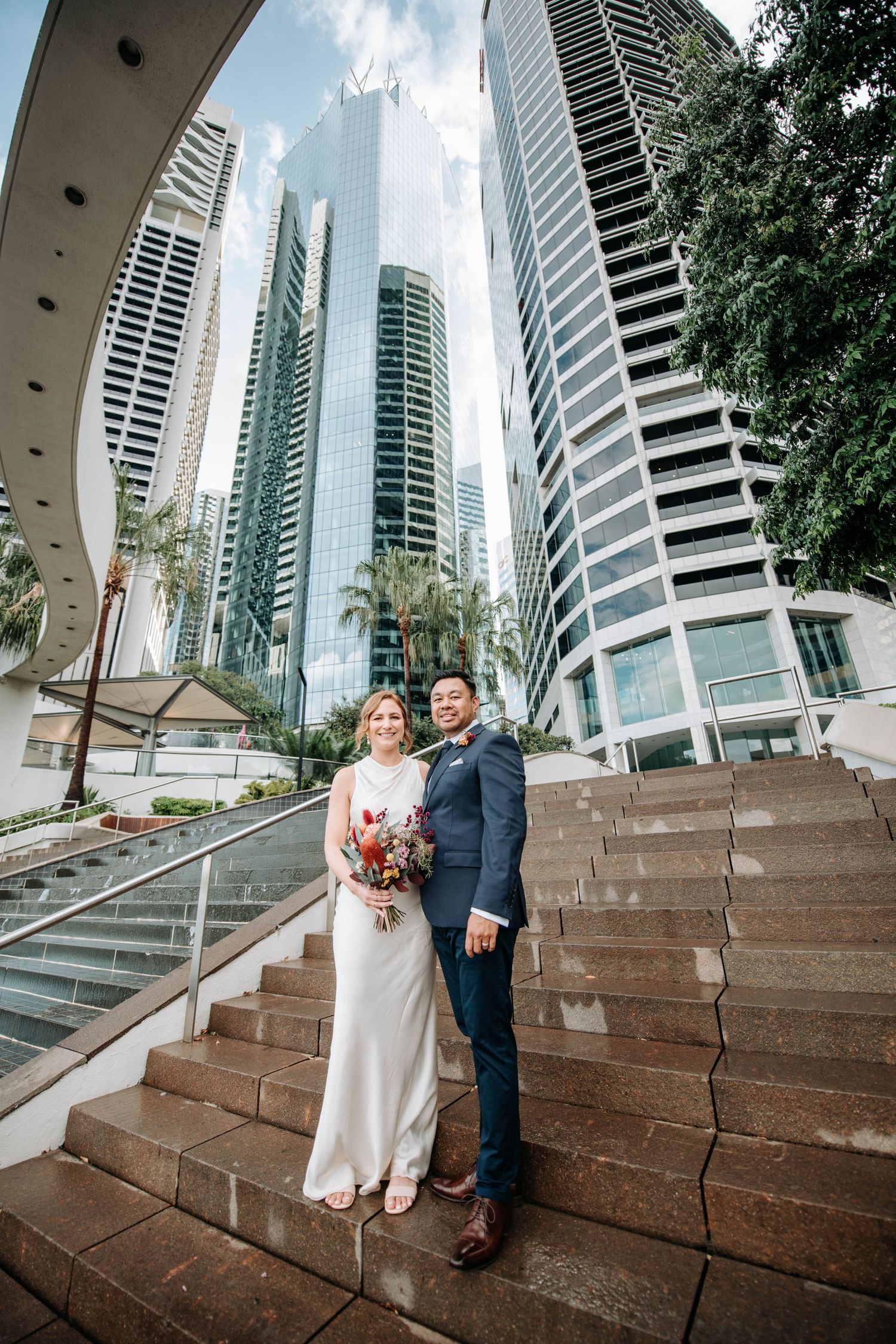 A wedding couple poses on stone steps against a backdrop of modern high-rise buildings and palm trees.