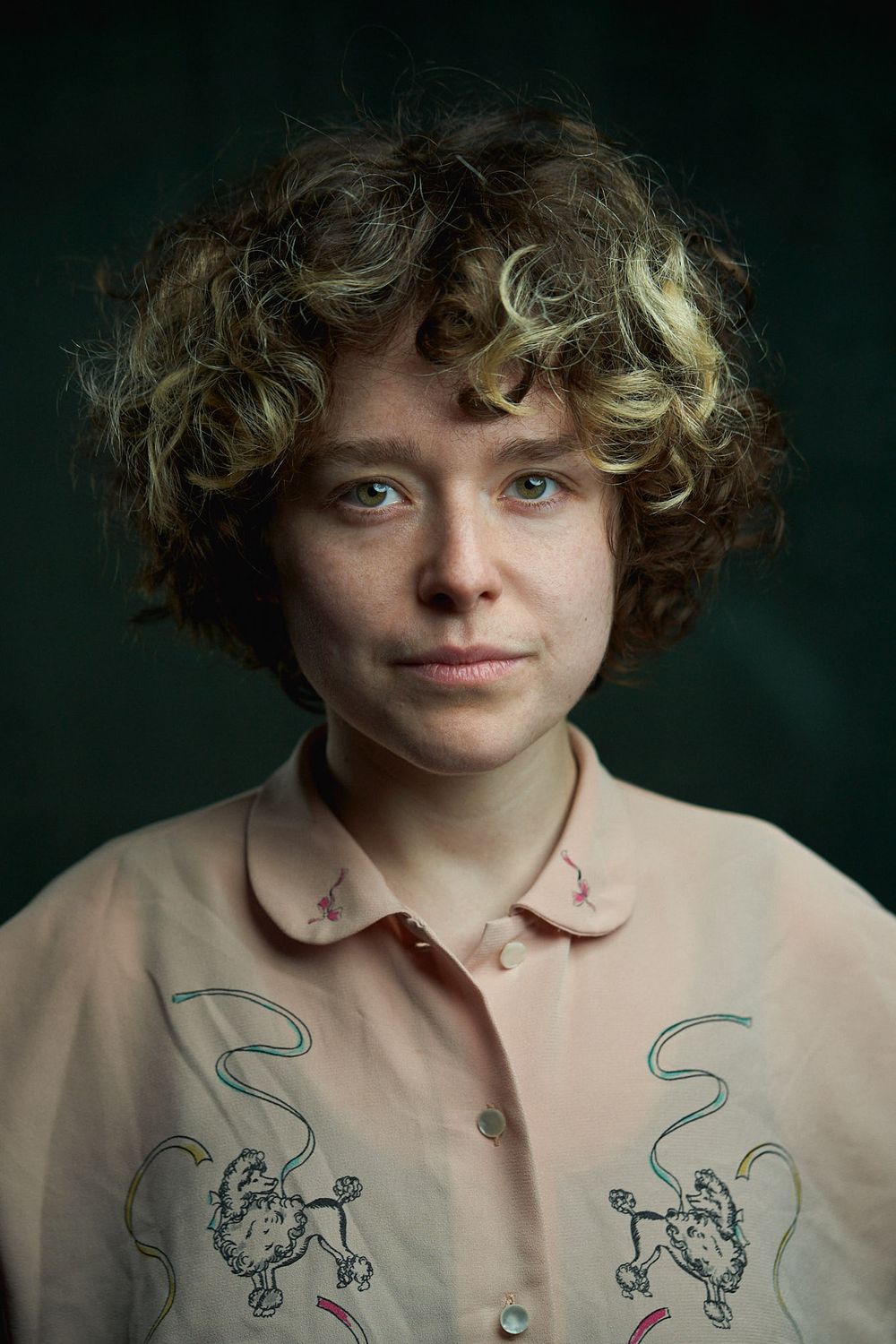 Portrait against dark green background wearing pink collared shirt with embroidered poodle designs.