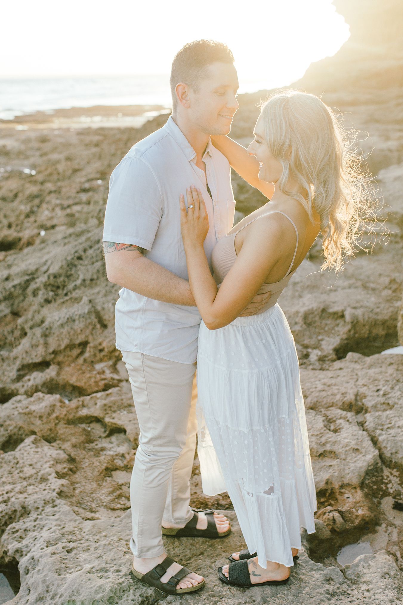 A romantic couple embraces on rocky coastal terrain during golden hour sunset.